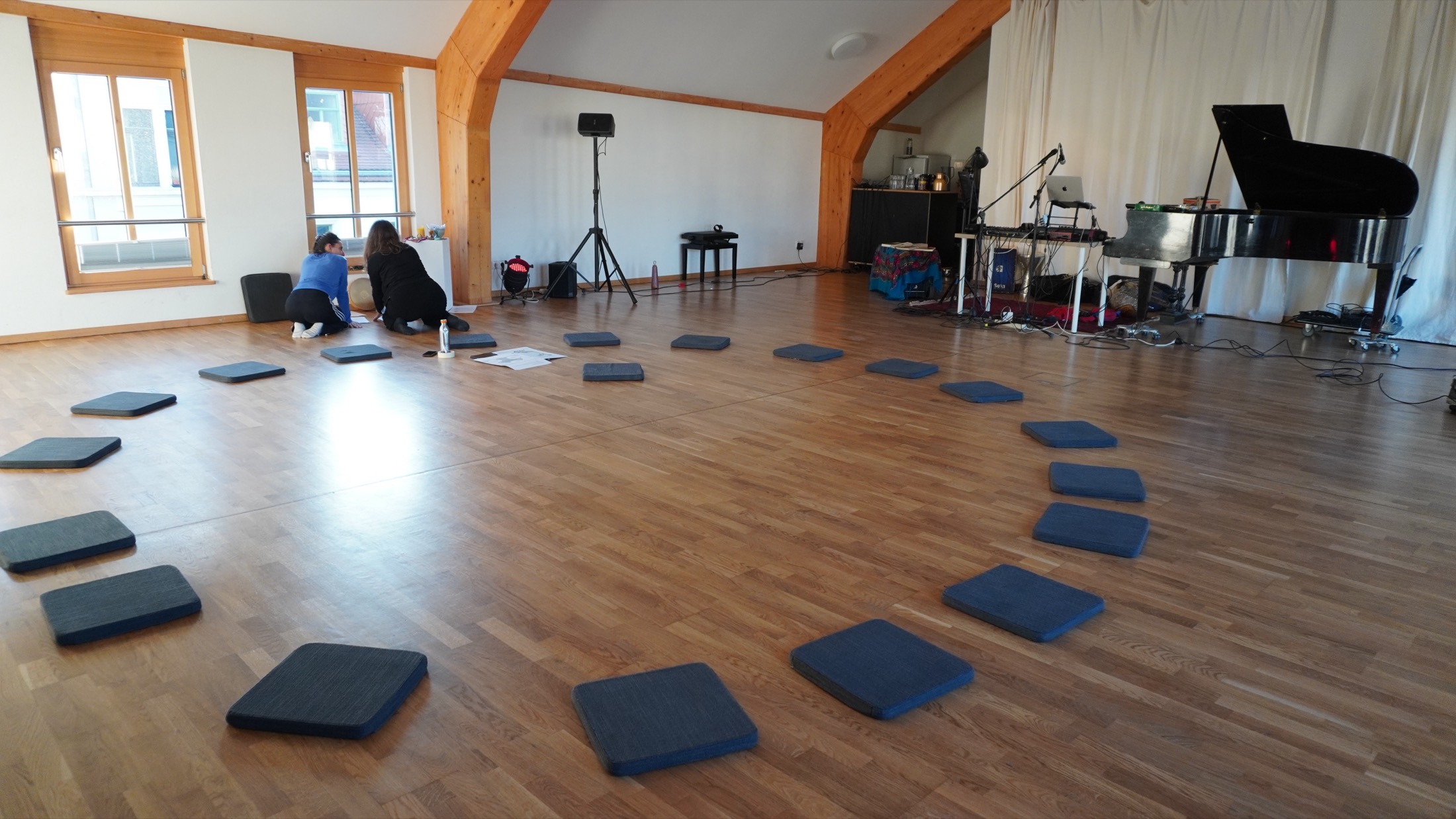 Floor cushions arranged in a wide circle in a bright hall with a wooden floor, a grand piano in the background